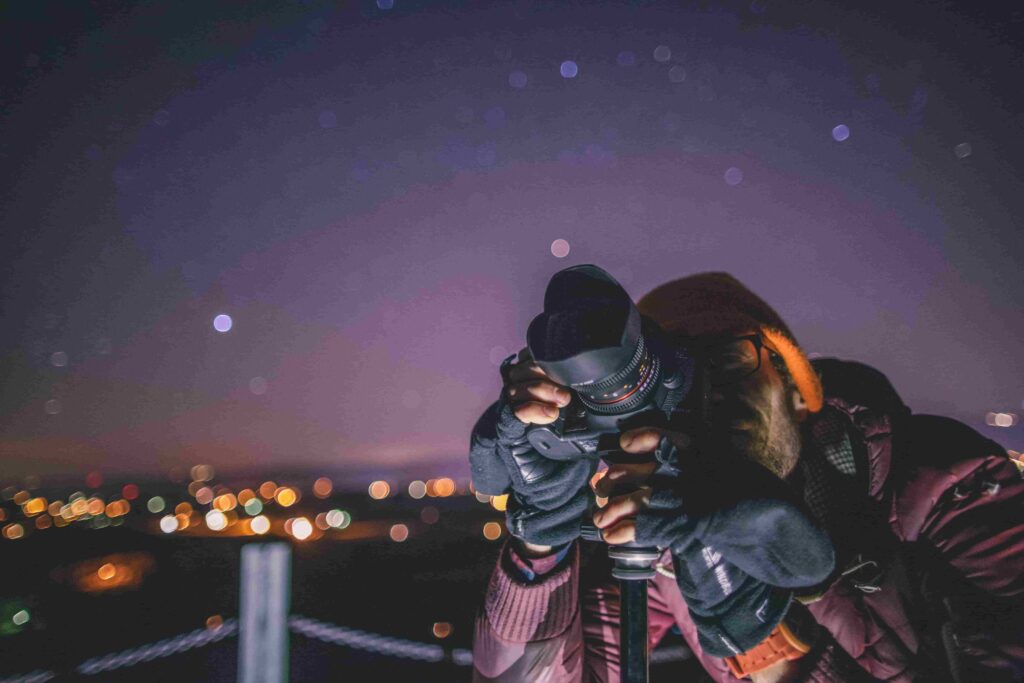 How to Photograph the Night Sky: A Complete Guide 1 - Blue Bend Photography Photographer shooting the night sky with a DSLR on a tripod with city lights bokeh in the background