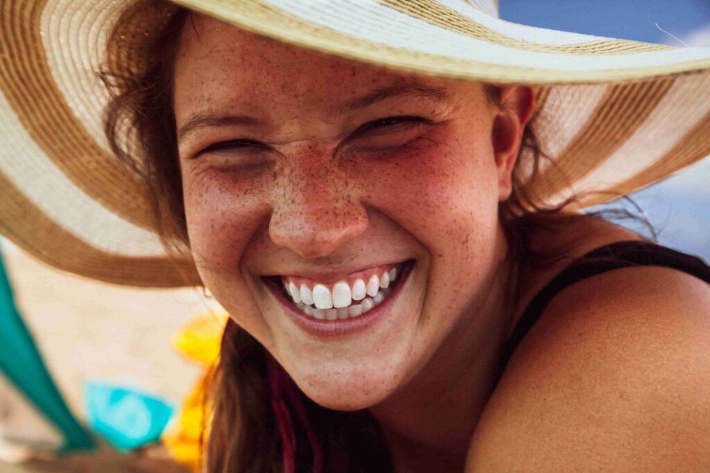 Portrait Photography Tips: Techniques, Settings & Lighting for Beginners 2 - Blue Bend Photography Close-up candid portrait of a laughing woman in a sun hat on the beach — natural expression portrait photography